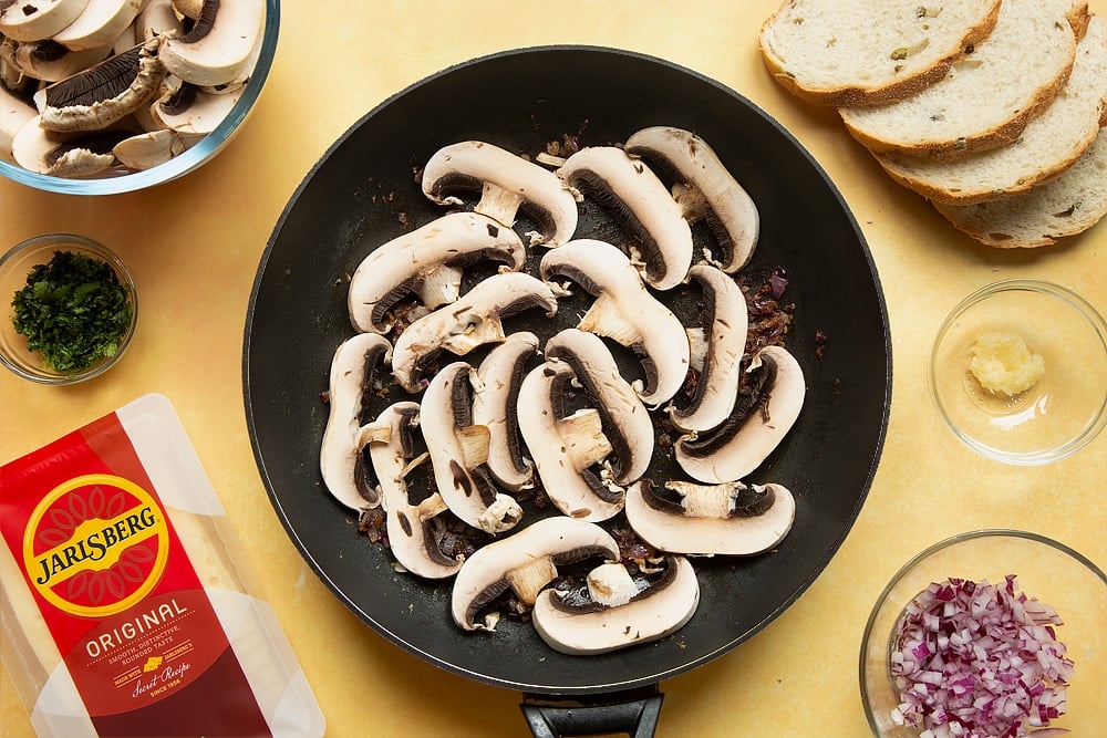 A frying pan with cooked garlic, onions, cayenne and black pepper in along with uncooked mushrooms in a single layer.