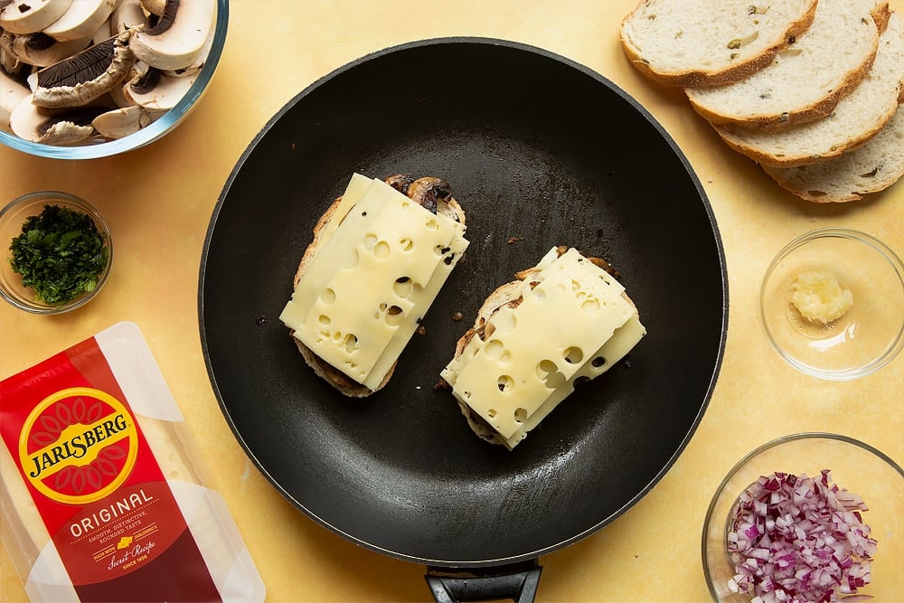 A frying pan with two slices of bread side by side and Jarlsberg® cheese on top, followed by cooked mushrooms, followed by more Jarlsberg cheese.
