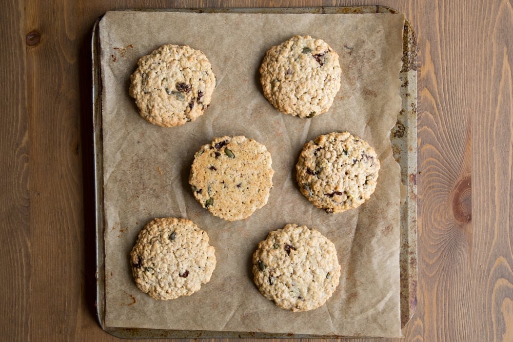 Freshly baked vegan oat cookies on a baking tray lined with baking paper. One is turned over to show the golden colour underneath.