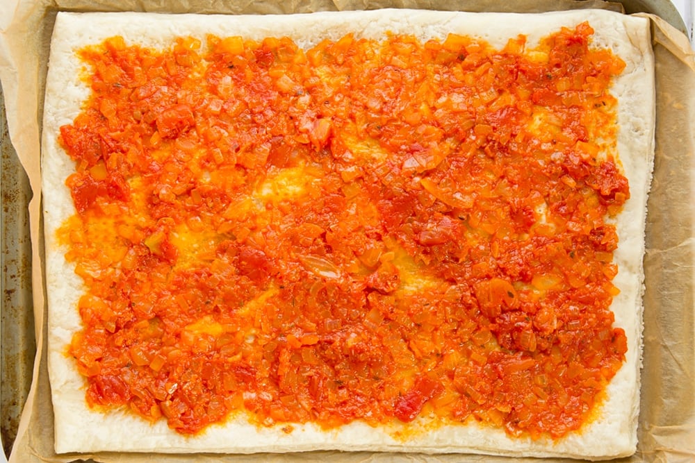 Overhead shot of puff pastry with spread out tomato sauce on a baking tray 