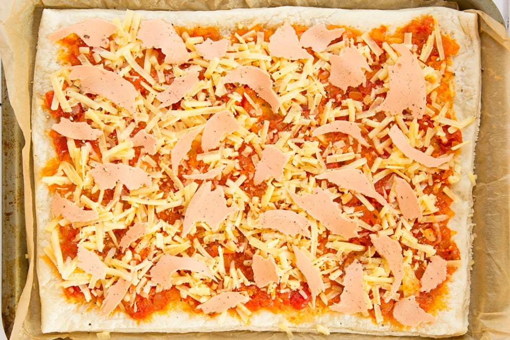 Overhead shot of puff pastry and tomato sauce topped with cheese and ham in a baking tray 
