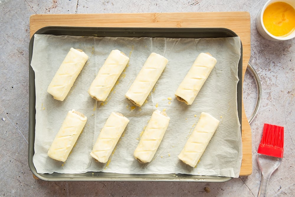 Eight vegetarian sausage rolls on a baking tray lined with baking paper. Each brushed with beaten egg.