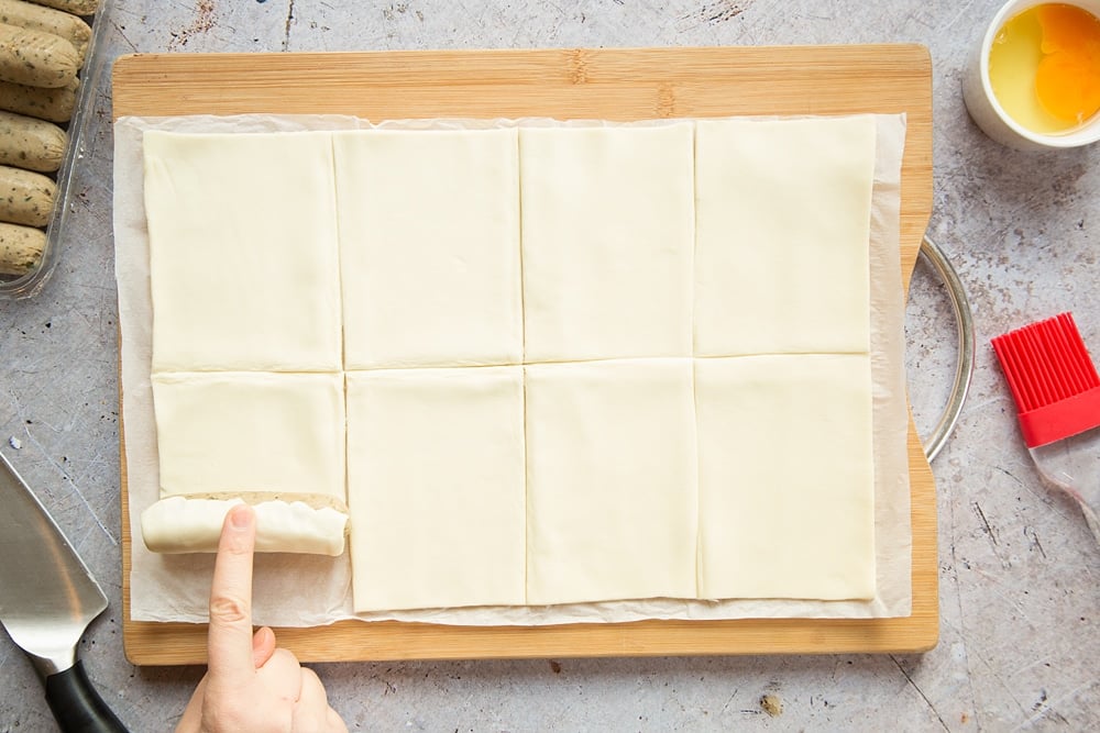 A sheet of puff pastry on a chopping board, cut into eight pieces, with a vegetarian sausage being rolled up in one piece,