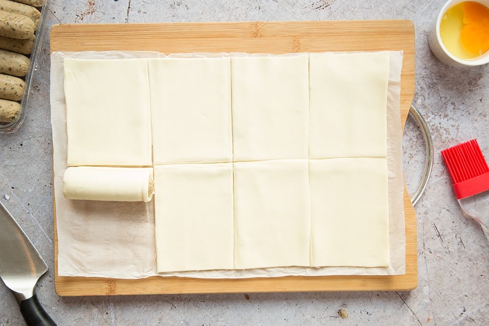 A sheet of puff pastry on a chopping board, cut into eight pieces, one vegetarian sausage fully rolled up in a piece of pastry.