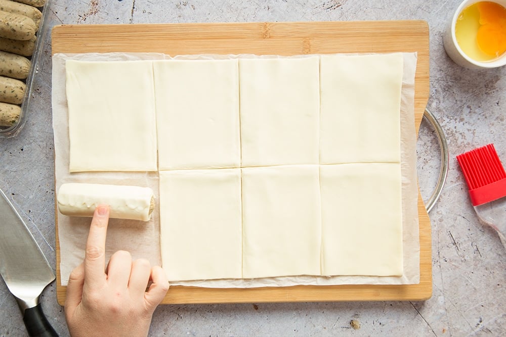 A sheet of puff pastry on a chopping board, cut into eight pieces, one vegetarian sausage fully rolled up in a piece of pastry. A finger pressing along the seam to seal.