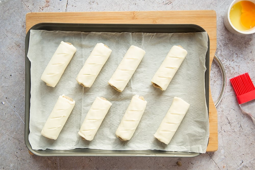 Eight vegetarian sausage rolls on a baking tray lined with baking paper. Three slits cut into the pastry of each one.