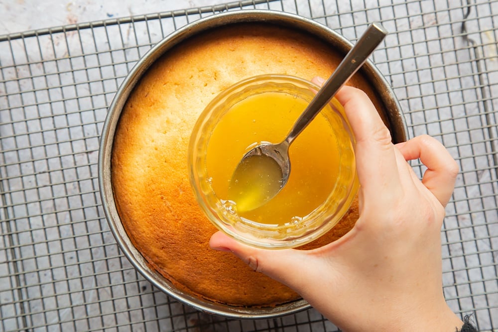 Holding a glass of lemon drizzle sauce over the cooled lemon drizzle cake.