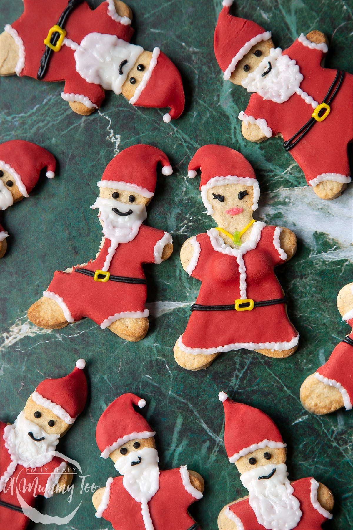 Lots of Father Christmas cookies on a green marble surface. One cookie has a bite take out of it. There is also a Mother Christmas cookie.