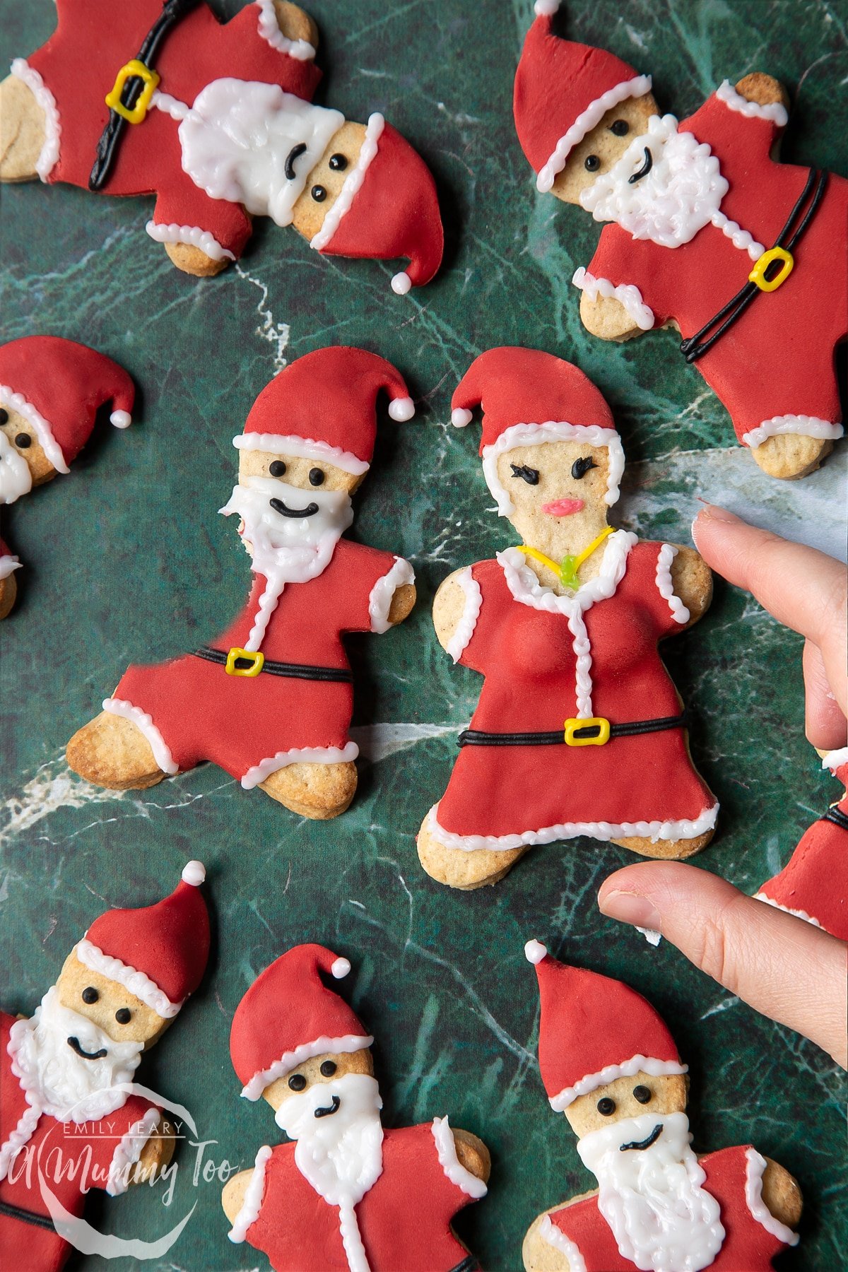 Lots of Father Christmas cookies on a green marble surface. One cookie has a bite take out of it. A hand reaches to take a Mother Christmas cookie.