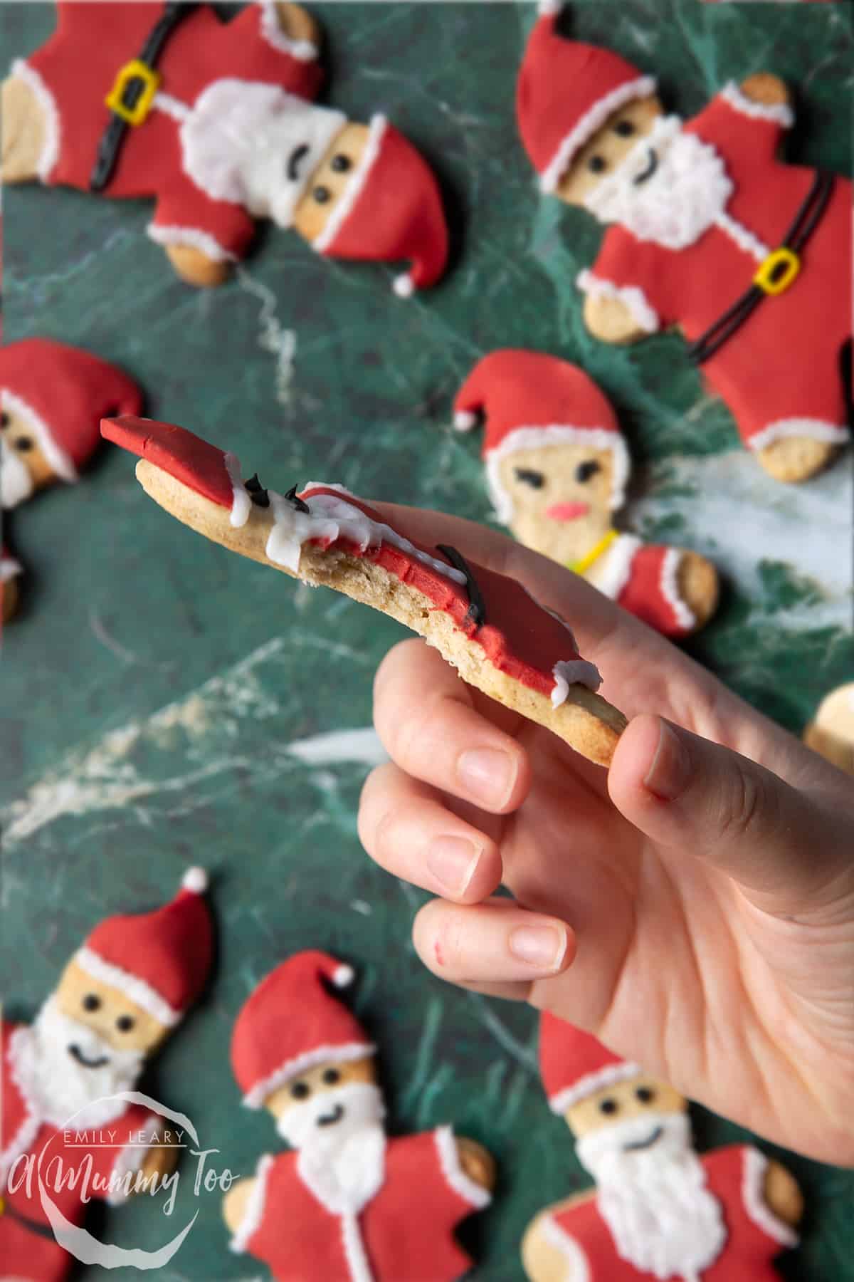 Hand holding a Father Christmas cookie on a green marble surface. A bite has been taken to reveal the crisp inner texture of the cookie.