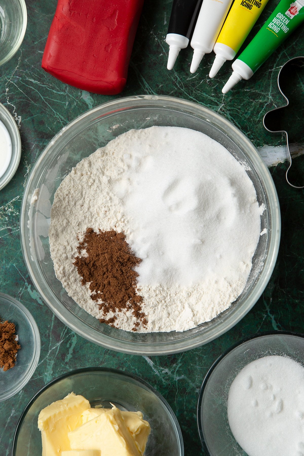 Flour, sugar and mixed spice in a glass mixing bowl, surrounded by ingredients to make Father Christmas cookies.