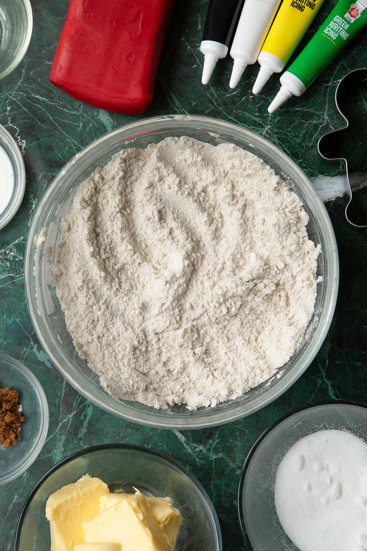 Flour, sugar and mixed spice combined in a glass mixing bowl, surrounded by ingredients to make Father Christmas cookies.