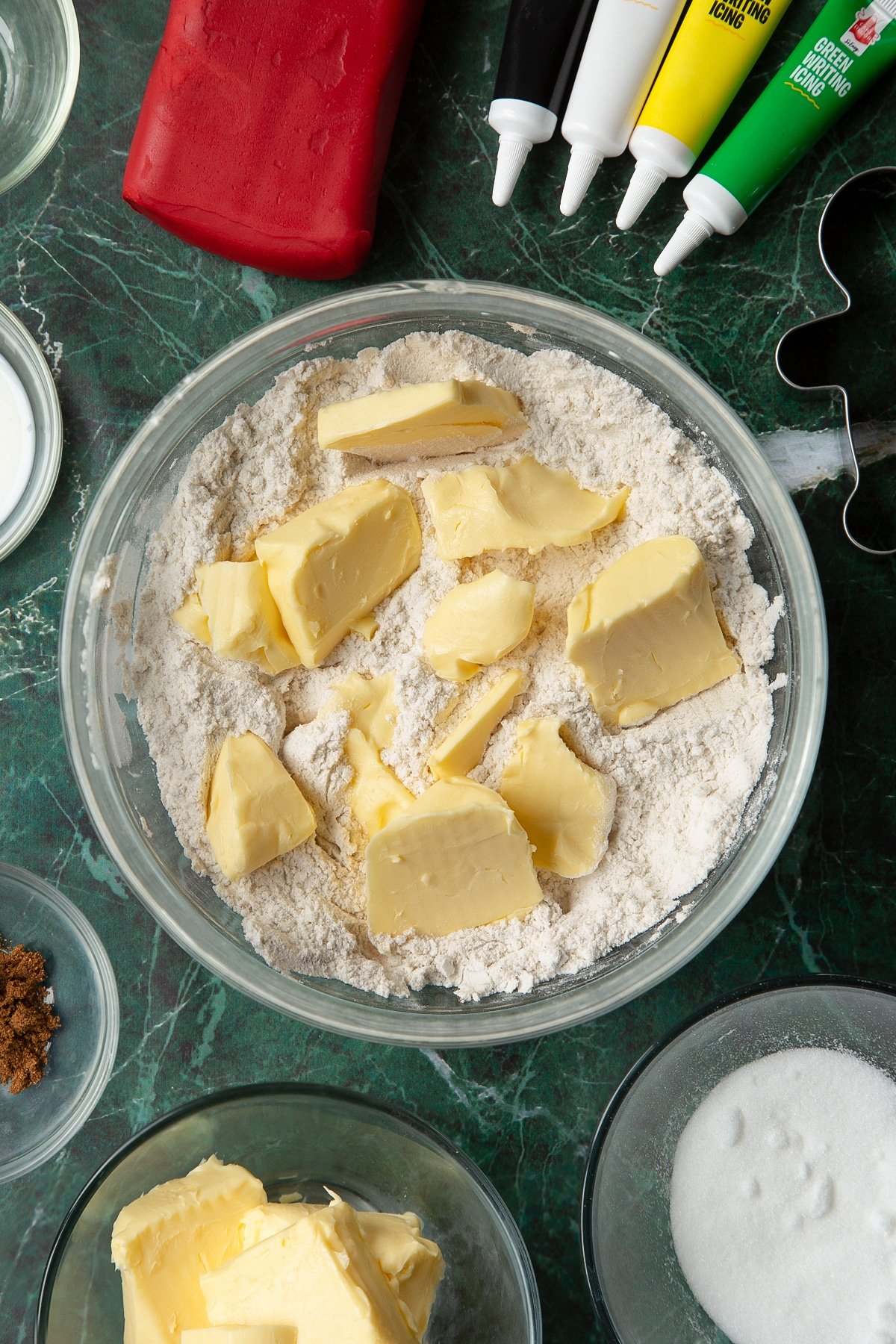 Flour, sugar and mixed spice combined in a glass mixing bowl with butter on top. The bowl is surrounded by ingredients to make Father Christmas cookies.