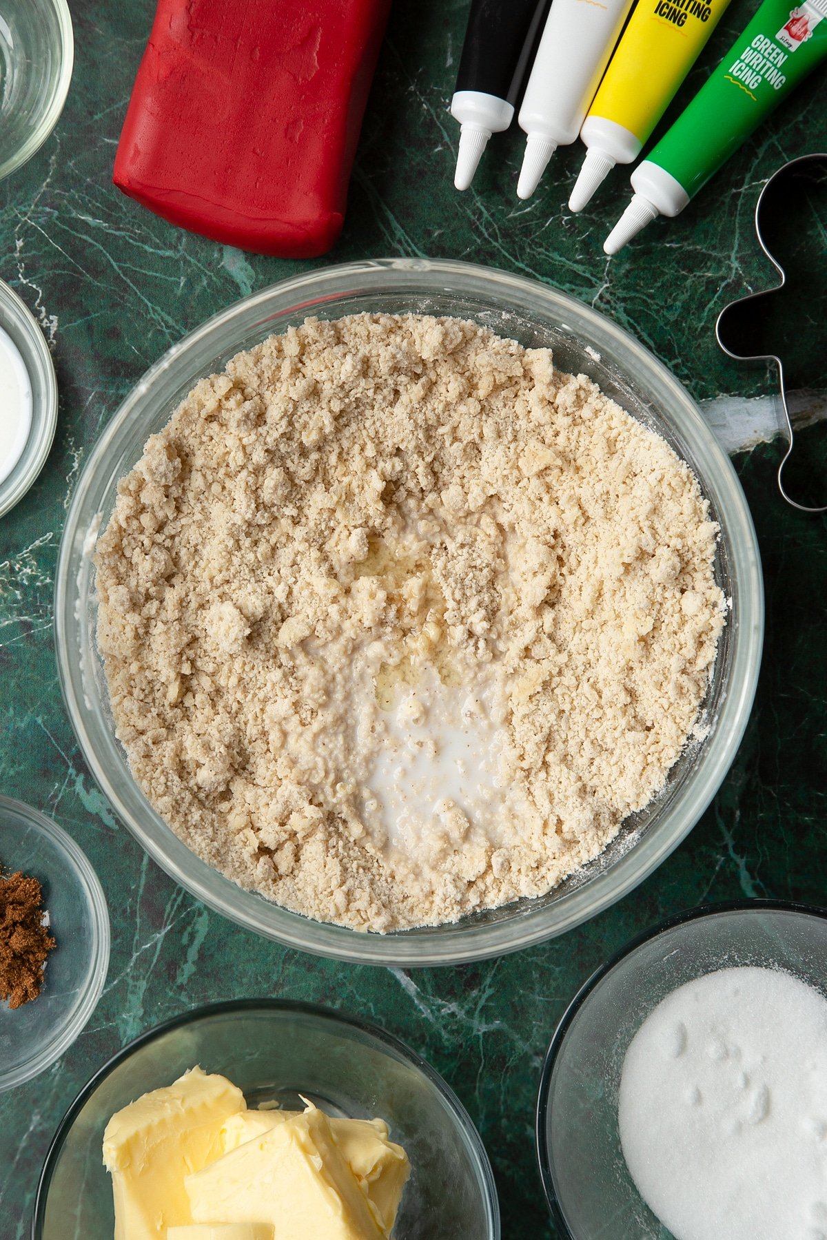 Flour, sugar, mixed spice rubbed together in a glass mixing bowl with milk & lemon extract on top. The bowl is surrounded by ingredients to make Father Christmas cookies.