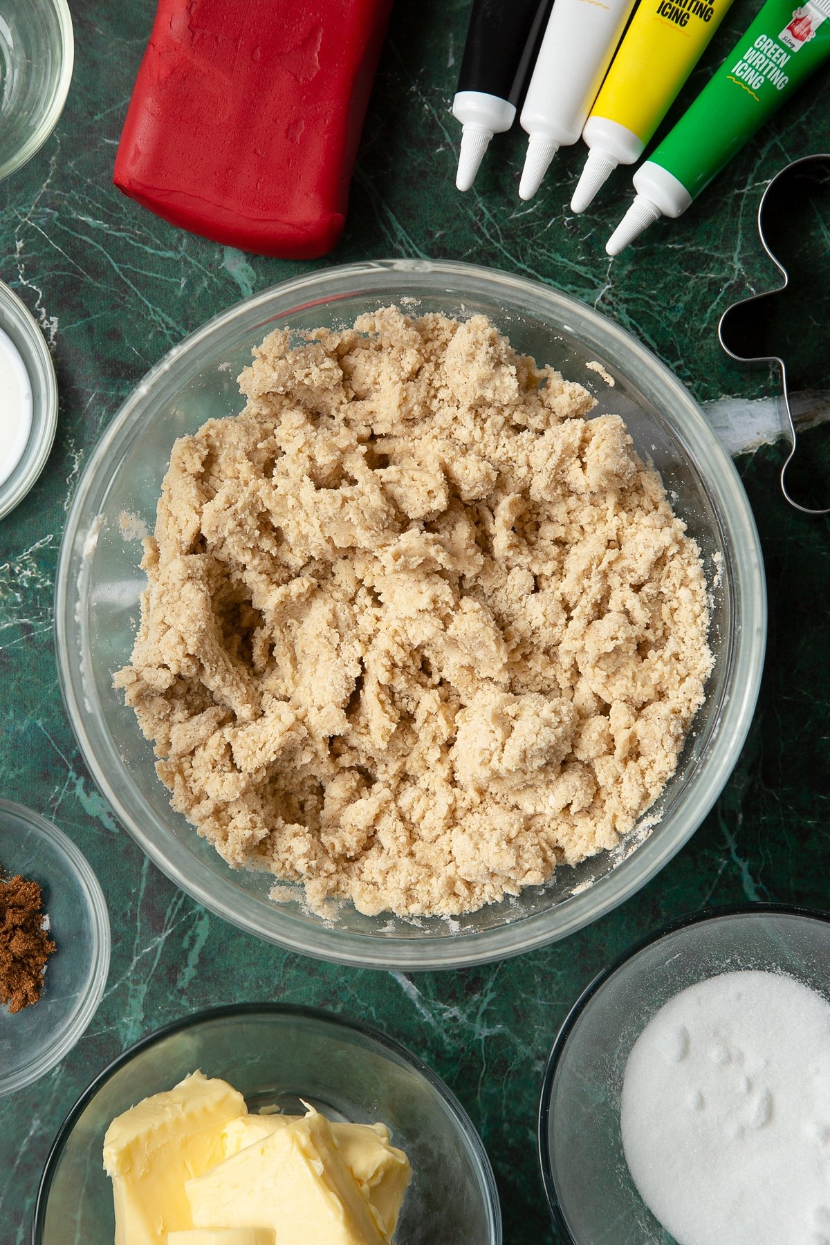 A rough cookie dough in a glass mixing bowl. The bowl is surrounded by ingredients to make Father Christmas cookies.