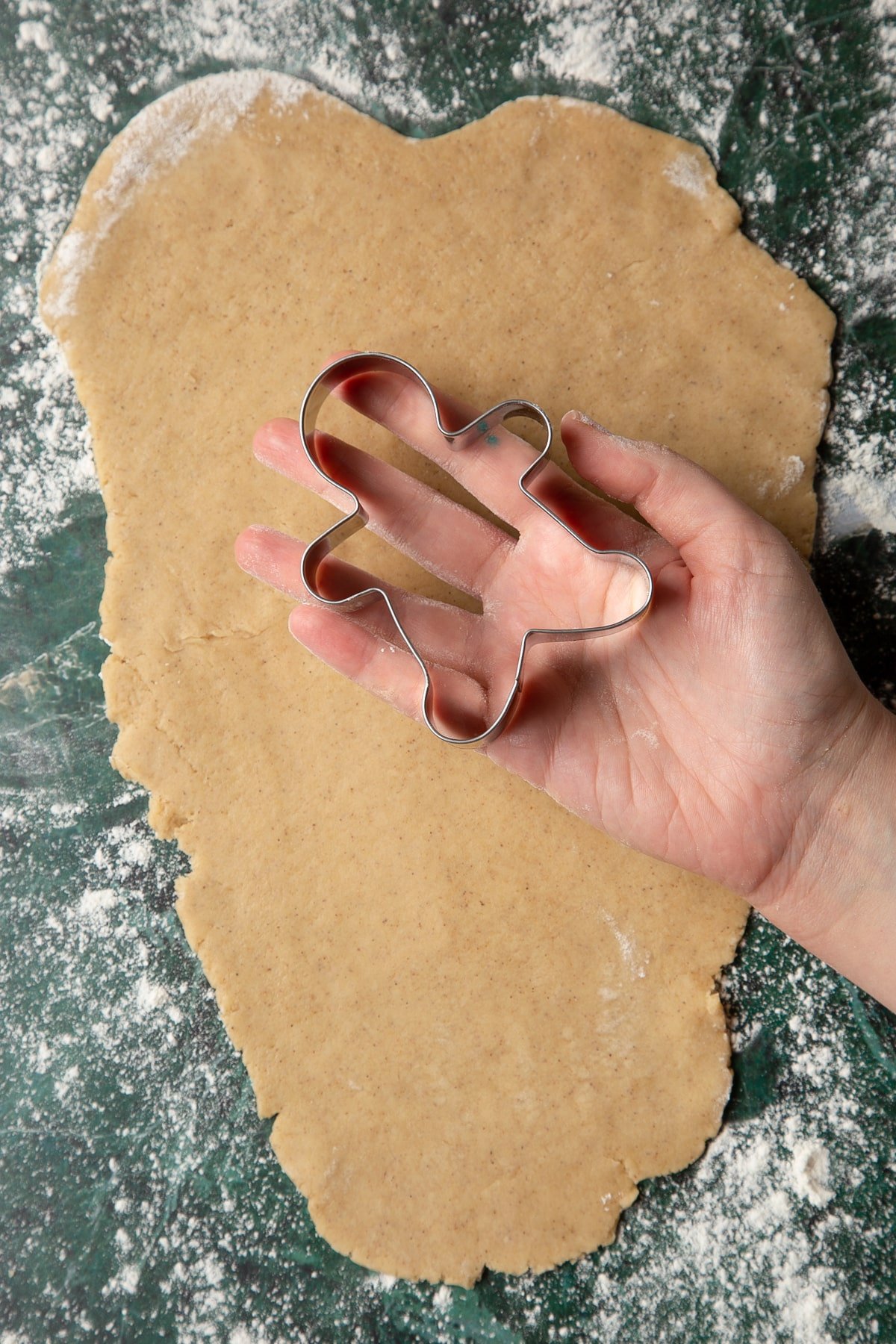 Hand holding a gingerbread man cutter about the size of the palm. Below it, cookie dough is rolled out.