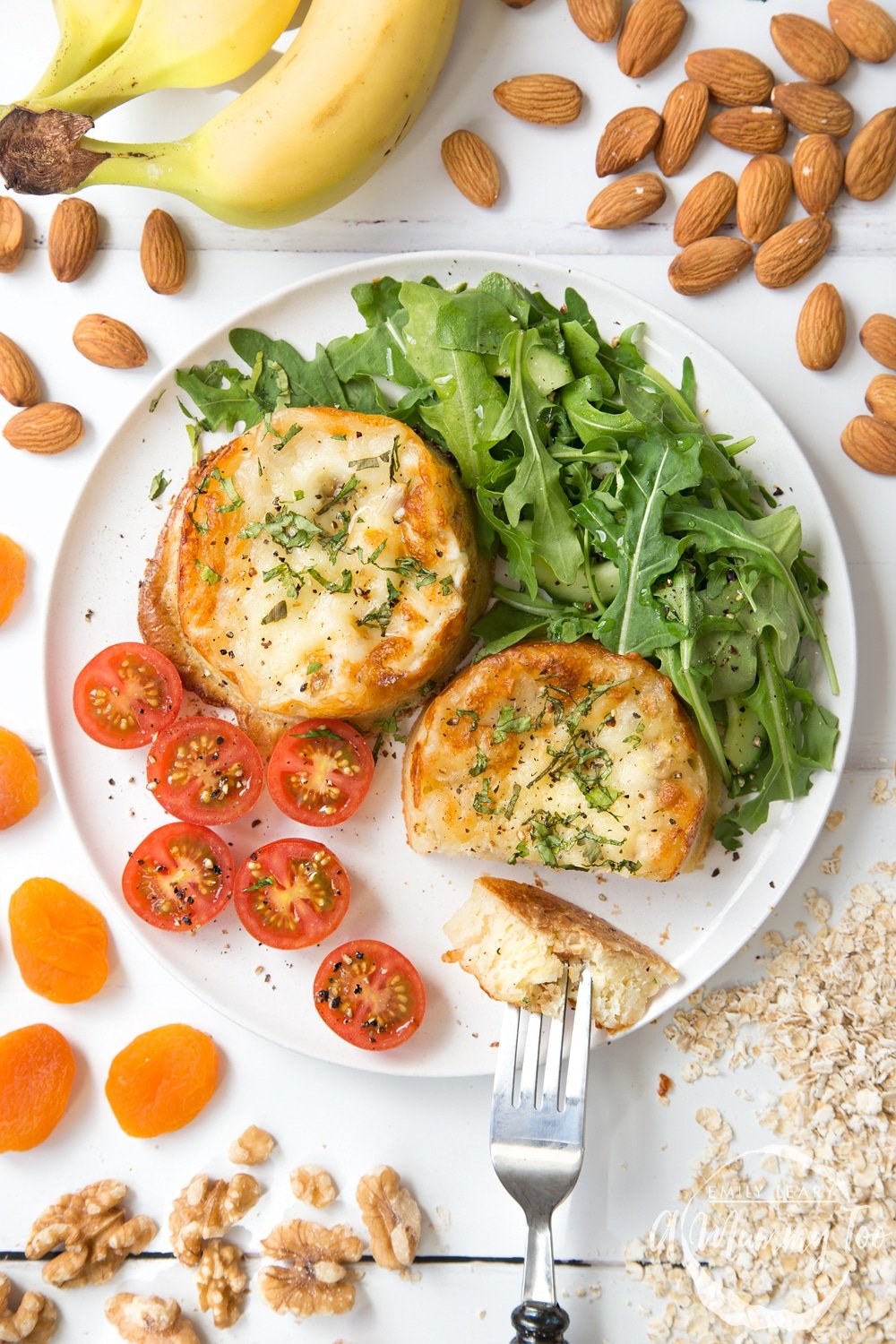 Overhead shot of the finishe Baked eggy crumpets on a white plate with a side salad. The plate sits on a white table with bananas and nuts on the side. 