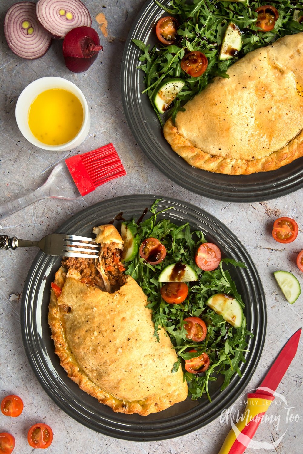 Meaty bolognese calzone with a side salad on a black plate surrounded by tomatoes, cucumber and onion.