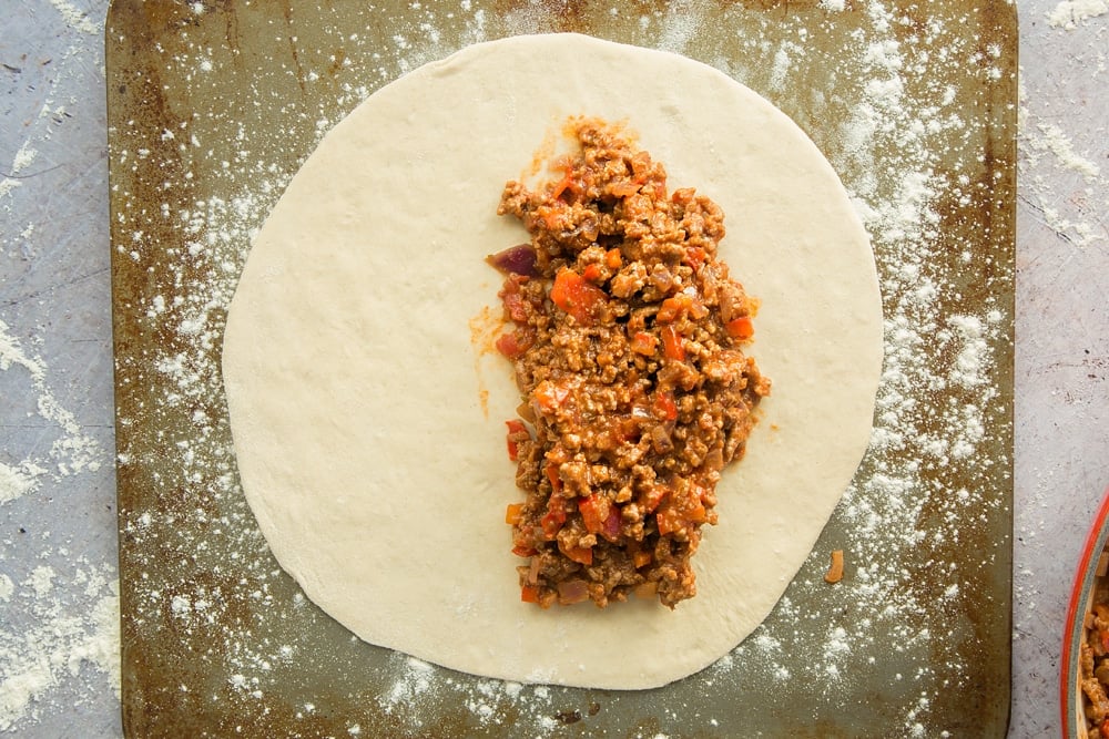 cooked mince meat mixture placed on one side of a large circular dough on a floured surface.