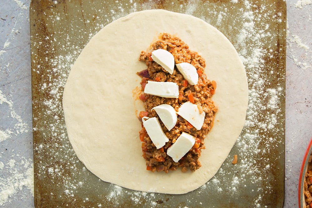 cooked mince meat mixture placed on one side of a large circular dough topped with mozzerella on a floured surface.