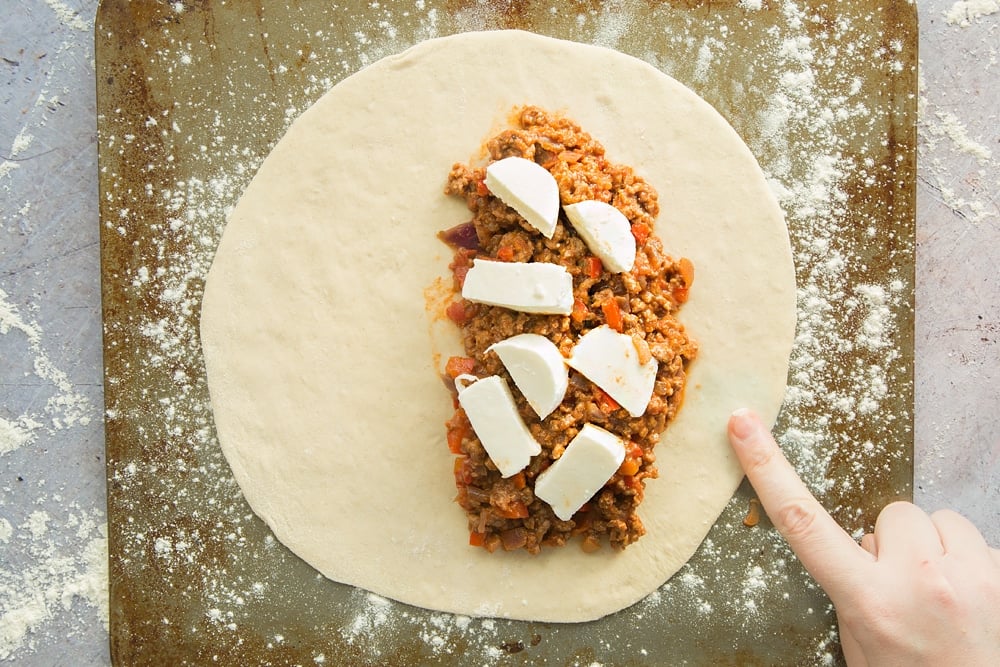 cooked mince meat mixture placed on one side of a large circular dough topped with mozzerella on a floured surface.