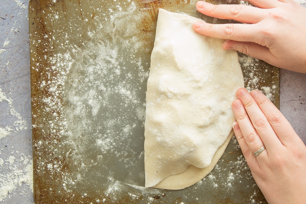 two hands folding over a floured dough to create a half circle encasing meat mixture.