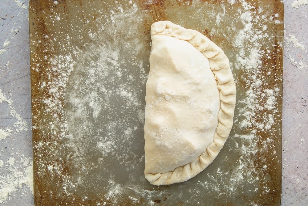 a half circle uncooked calzone pizza on a floured baking tray.