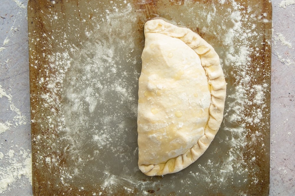 a half circle uncooked calzone pizza on a floured baking tray topped with sea salt and oil.