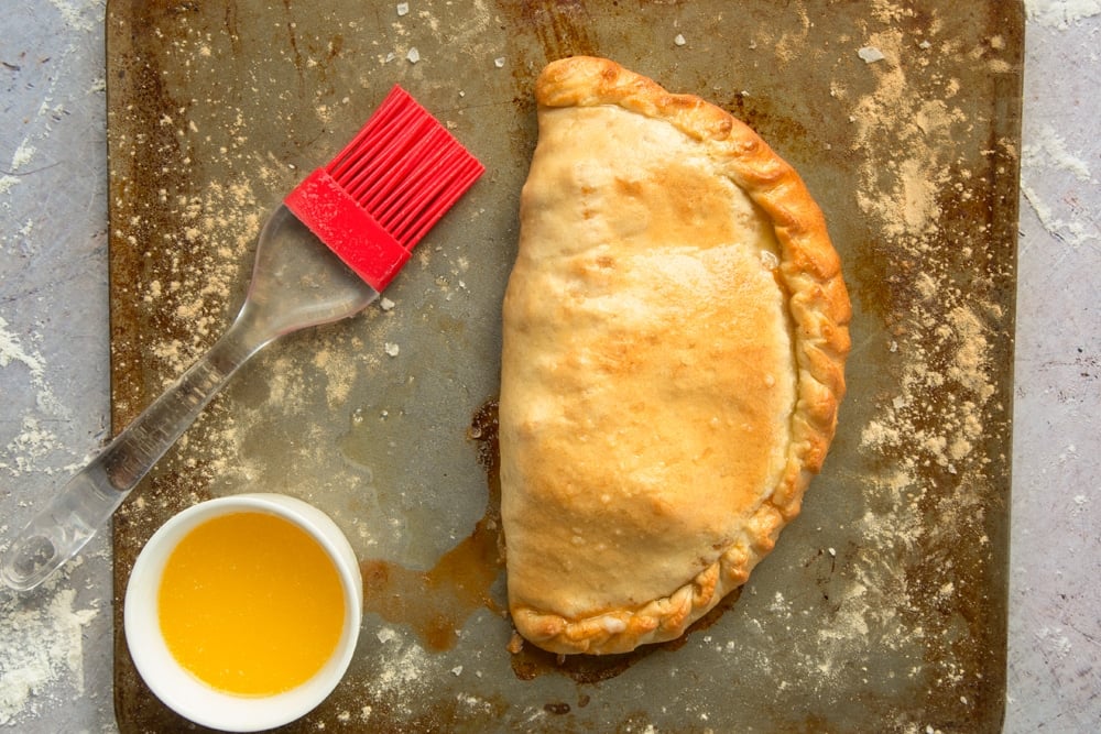 a half circle cooked calzone pizza on a floured baking tray with a pot of oil and brush on the side.