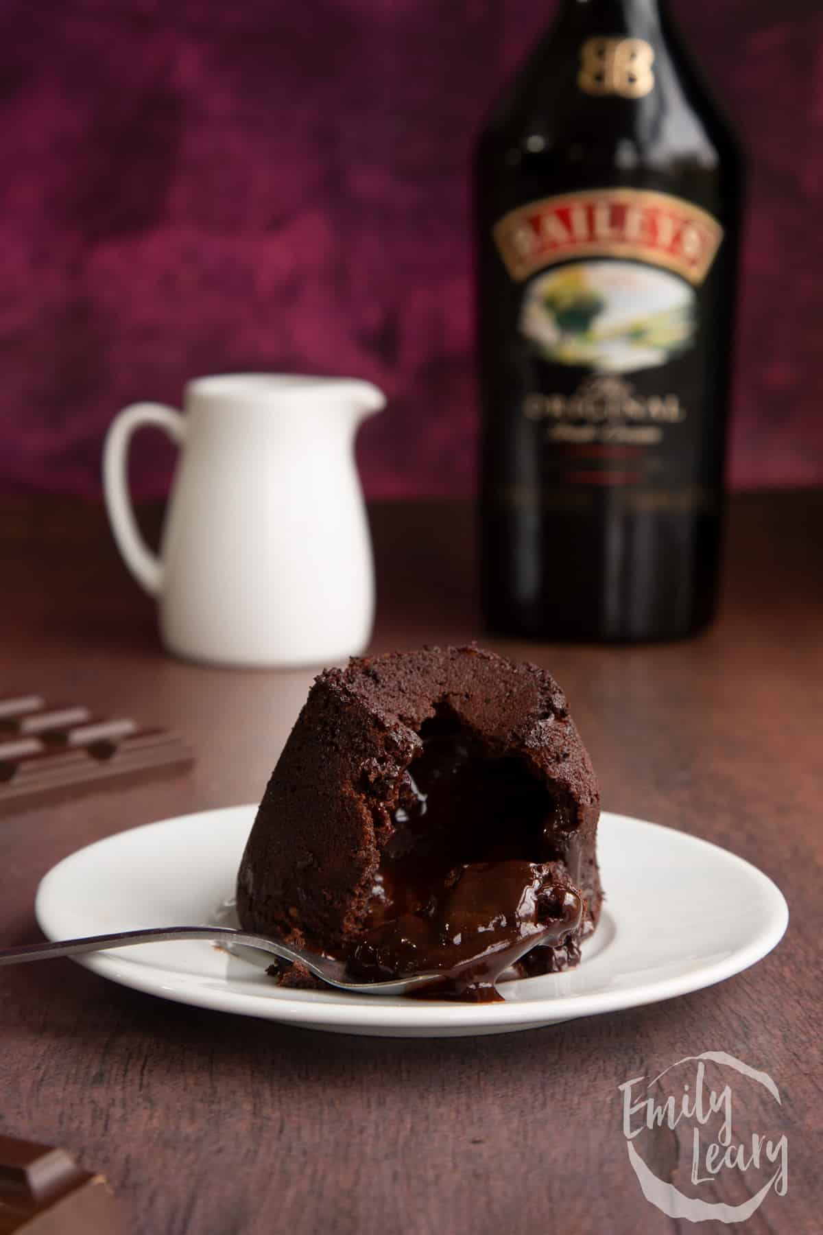 Baileys lava cake on a white plate with a bottle of baileys in the background cut open by a spoon.