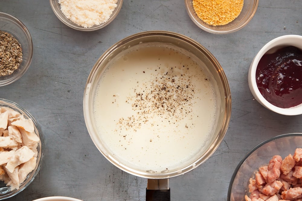 Preparing white sauce for the festive slice. The ingredients are currently in a pan. Additional ingredients for the recipe are on a dark grey background in bowls. 
