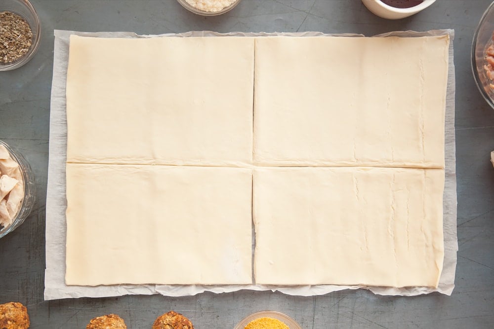 The pastry having been rolled out and cut evenly into four rectangles on a dark grey background with bowls of additional ingredients for the recipe at the side.