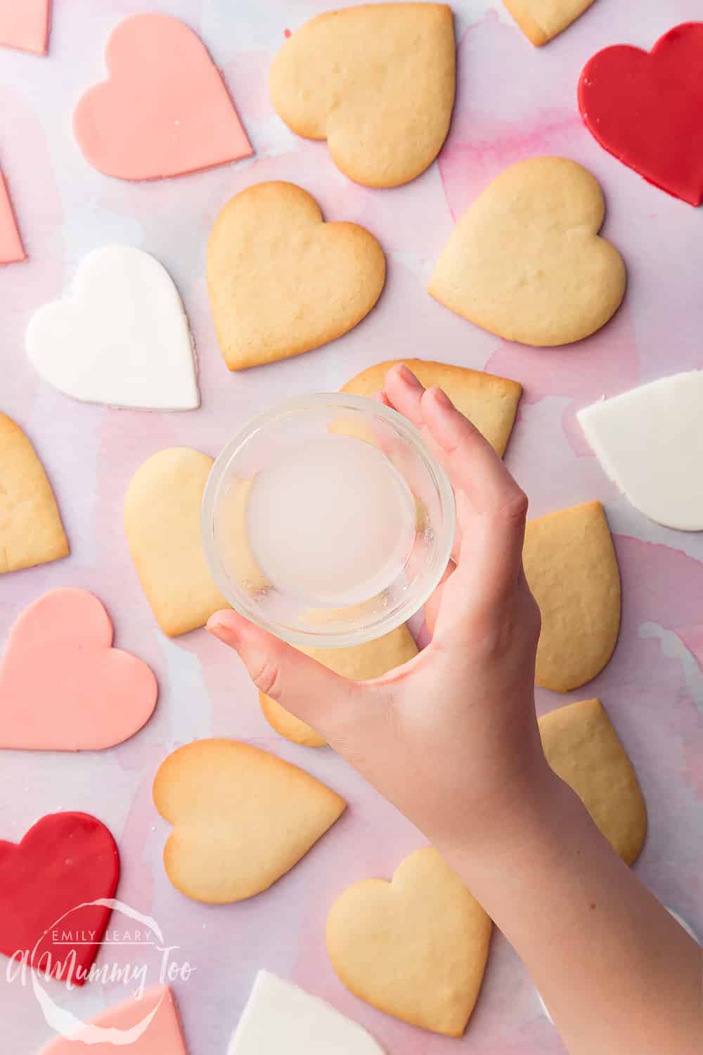 Overhead shot of a hand holding a small clear dish with icing sugar over Heart biscuit for Valentine's Day with a mummy too logo in the lower-right corner