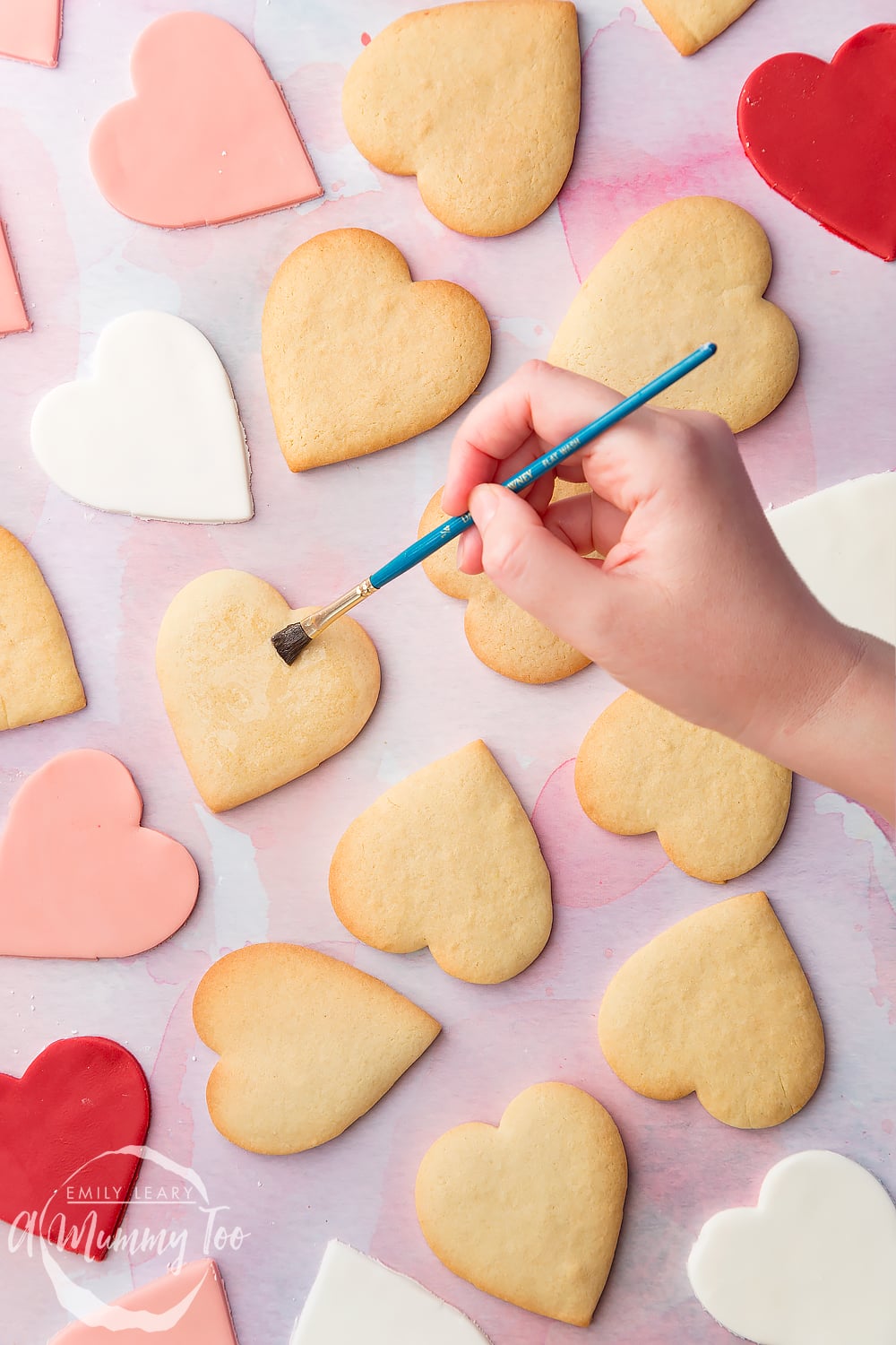 Overhead shot of a hand holding a brush and glazing heart shaped biscuits with icing sugar with the A Mummy Too logo in the bottom left hand corner