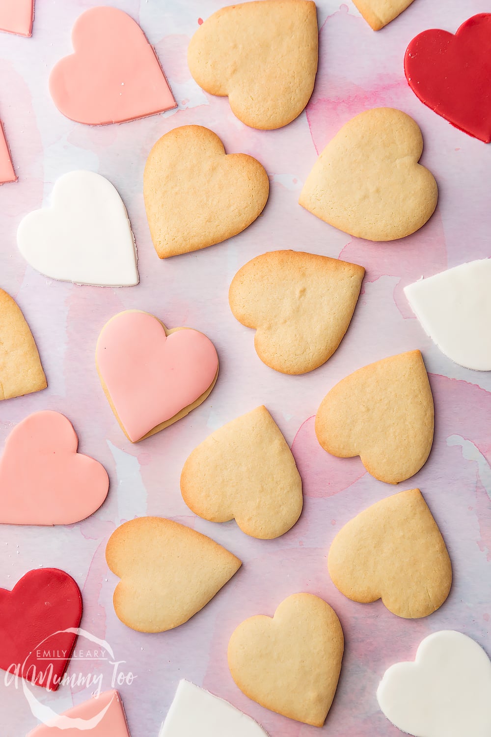 Overhead shot of heart shaped biscuits with some topped with pink fondant with the A Mummy Too logo in the bottom left hand corner