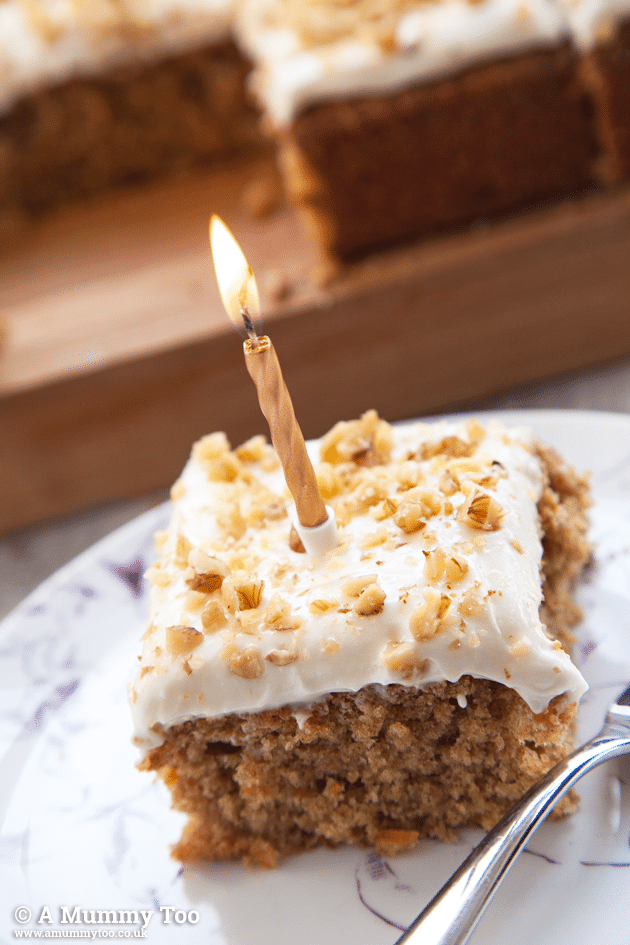 square piece of carrot cake topped with frosting and nuts with a fork on the side and a candle in the top.