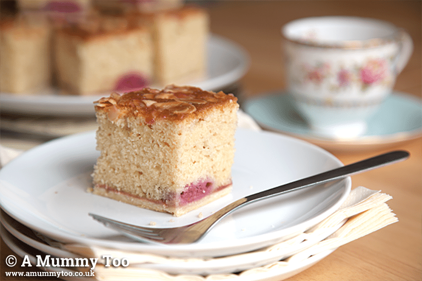 a piece of Raspberry Bakewell Tray Bake on a small white plate with a fork with a teacup in the background