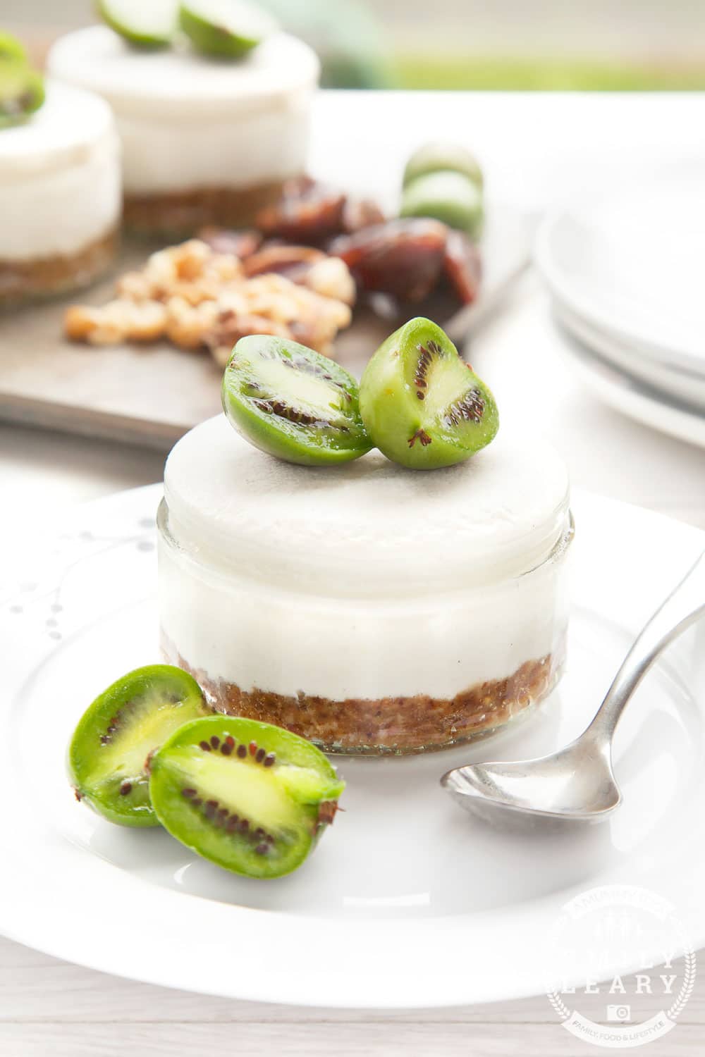 Mini kiwi berry cheesecakes in clear small jar with kiwi berries and a spoon and more mini cheesecakes in background.