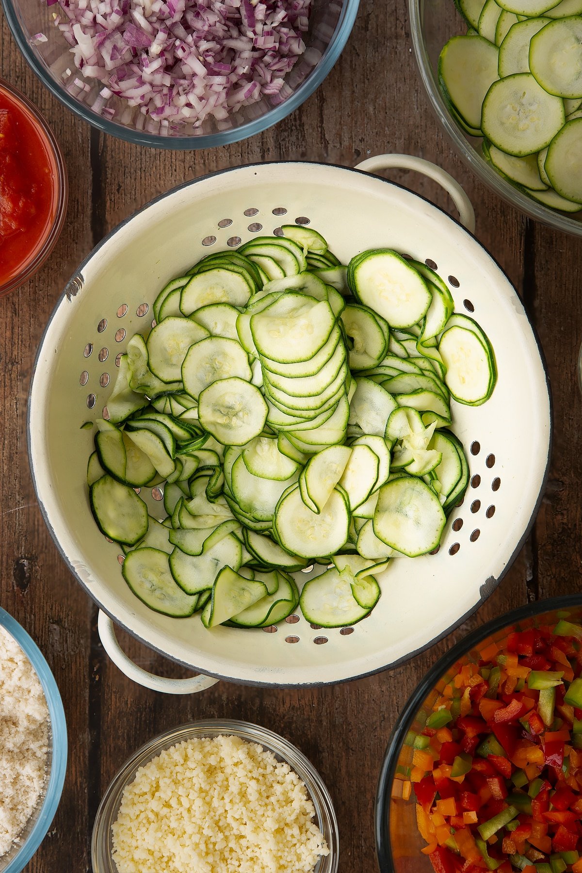 thinly sliced courgette in a white colander with ingredients around the edges.