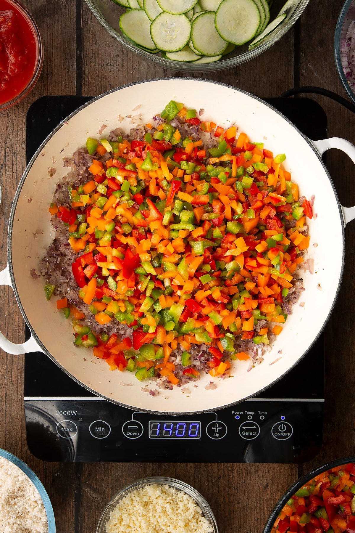 cooked dred onion and garlic mix topped with diced peppers in a large white frying pan on induction hob.
