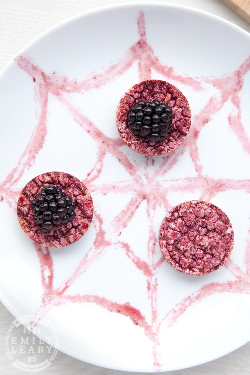 A white plate decorated with a spider web pattern using blackberry juice. Rice cakes and blackberries have been added. 