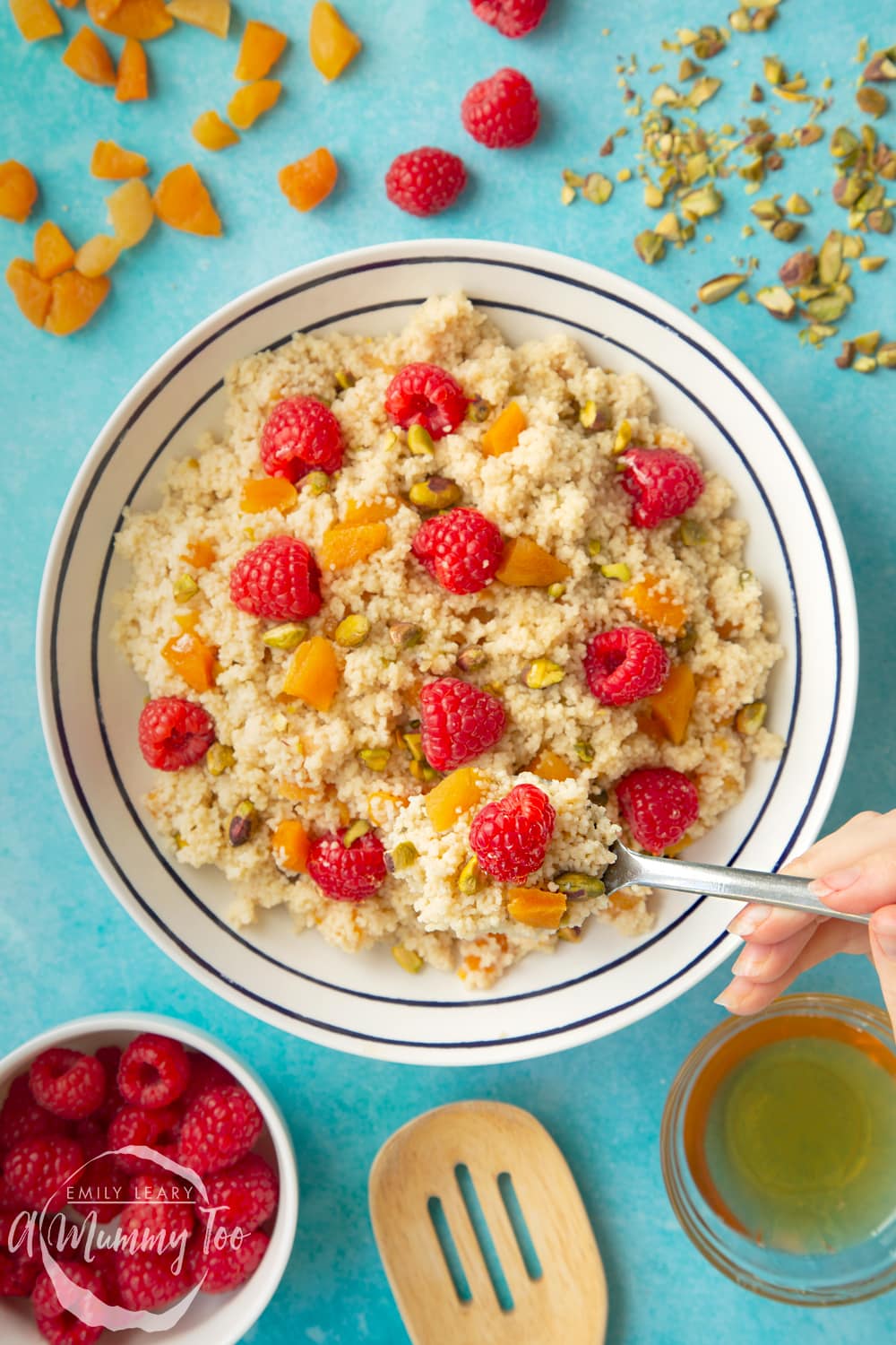 Overhead shot of a spoon going into the bowl of sweet breakfast couscous. The bowl is surrounded by ingredients required for the recipe. 