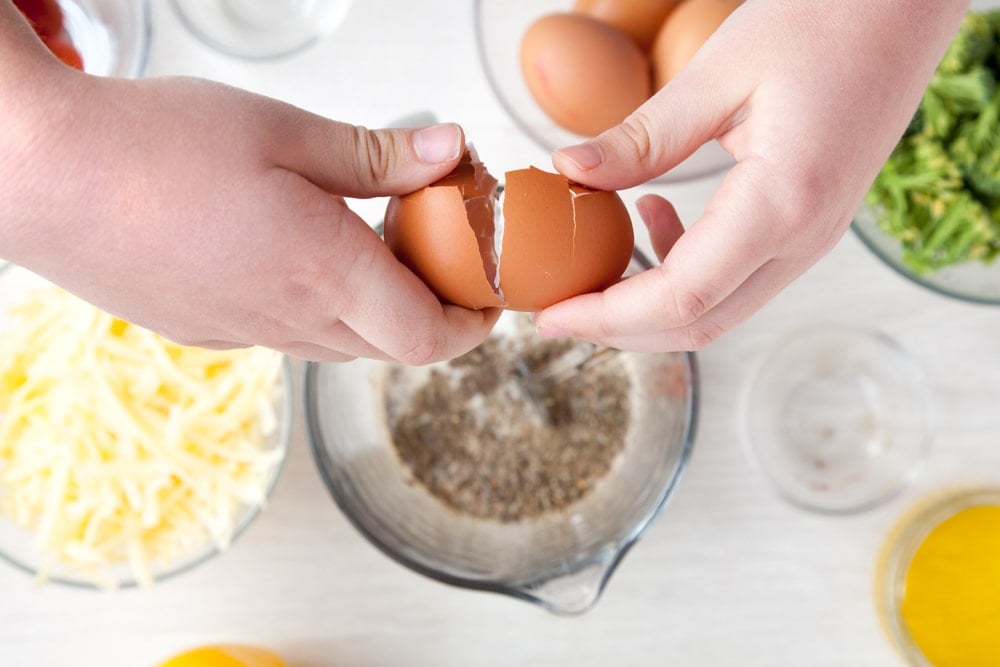 Breaking eggs into the quiche mix which is currently in a jug on a white wooden table.