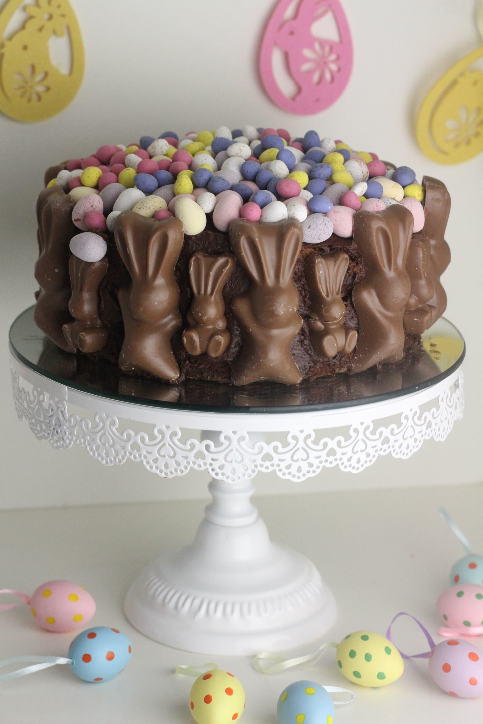 Malt Easter Chocolate Cake topped with mini eggs and trimmed with malteaser bunnies served on a white metal cake stand.