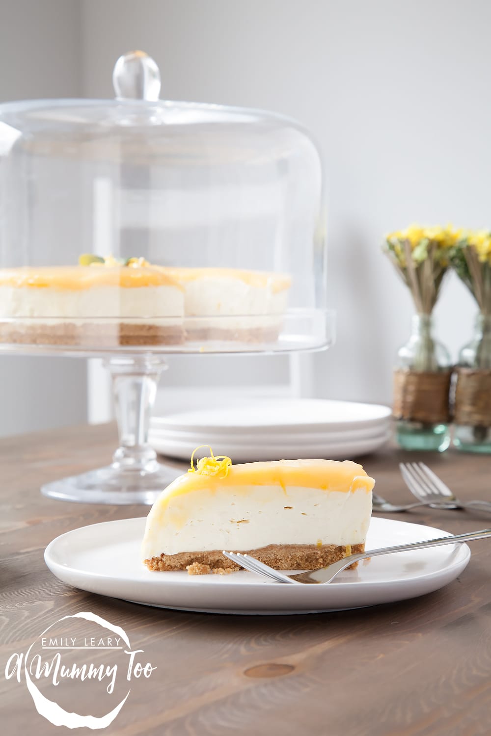 A slice of no-bake lemon curd and white chocolate cheesecake on a plate, with the remaining cheesecake in a stand behind.