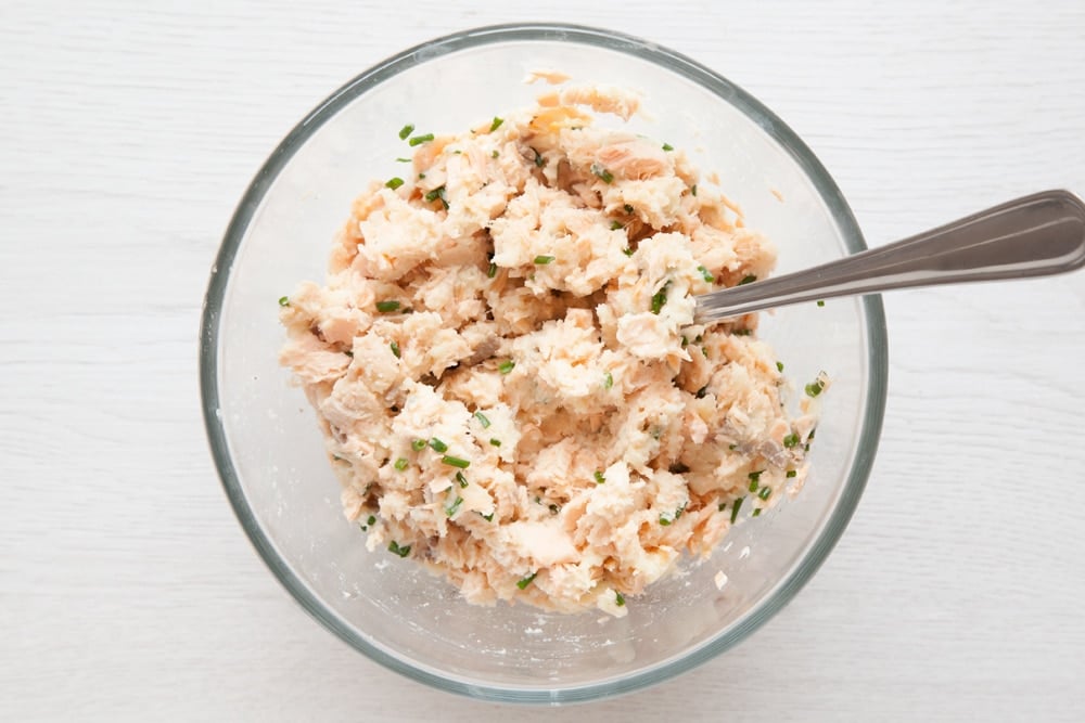Salmon fishcake mix in a bowl with a fork on the side. The bowl is set on a light wooden background. 