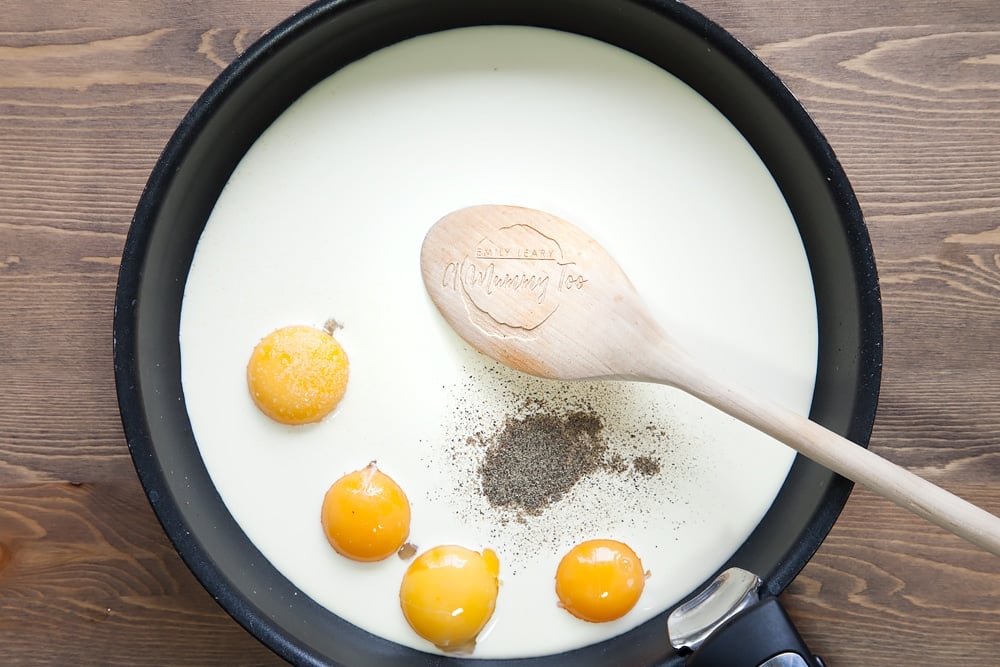 Cream and 4 egg yolks in a frying pan accompanied by a wooden spoon, with salt and pepper