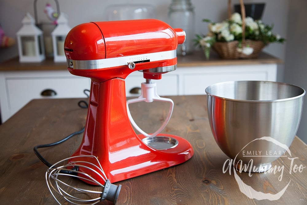 KitchenAid Mini in red sat on a wooden table. At the side there are some of the accessories for the appliance. 