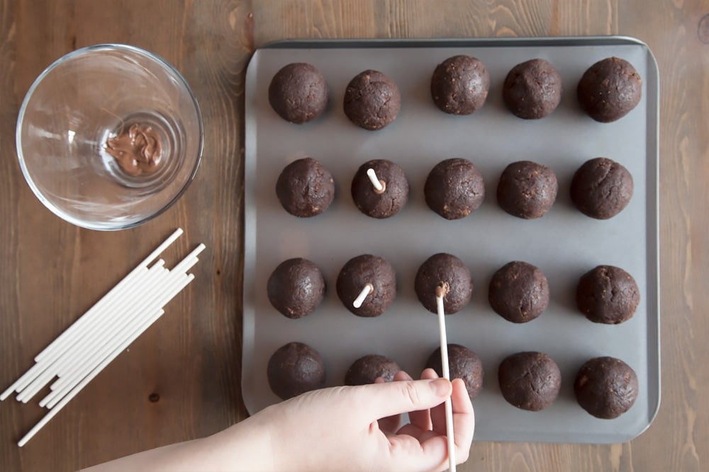 Bowl of melted chocolate with some cake pop sticks being pressed into the balls of cake mixture for the kitty cake pops.