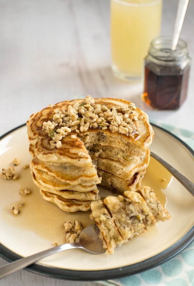 Granola pancakes by amuse your bouche tsacked on a white plate topped with granola and syrup with a bit missing from the side.